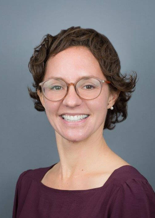 Smiling woman with short brown hair and glasses, wearing a purple top, posed against a gray backdrop.